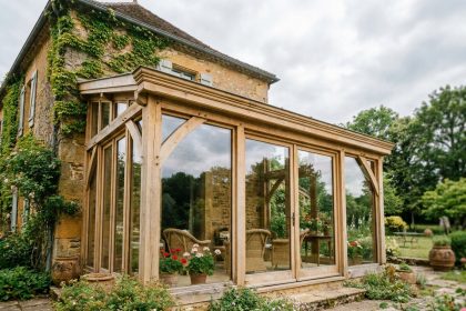 Verrière en bois sur une maison familiale vue depuis le jardin, structure en chêne naturel et panneaux vitrés intégrés à une façade en pierre